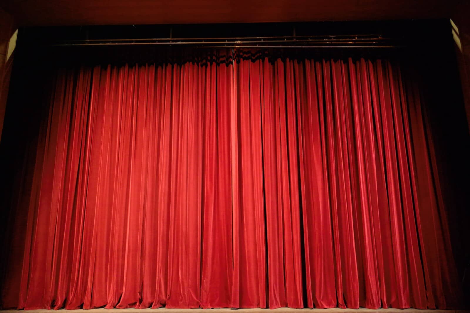 A large stage with closed, deep red velvet curtains, evenly pleated from top to bottom, under dim theatre lighting.