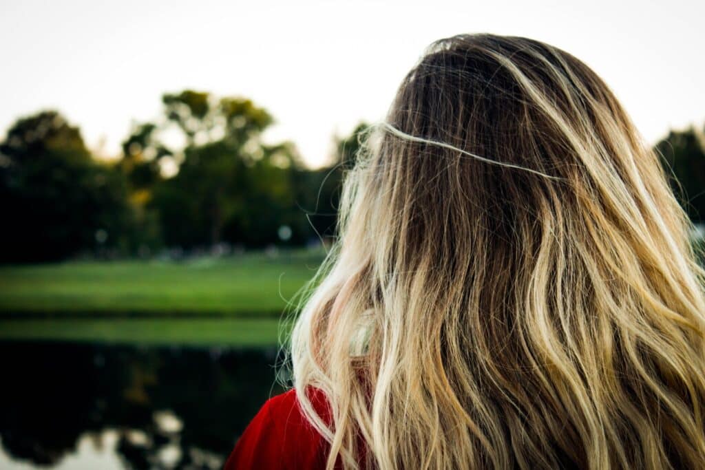 A person with long, wavy blonde hair stands outdoors, facing away from the camera, with a grassy area, trees, and a body of water visible in the background.