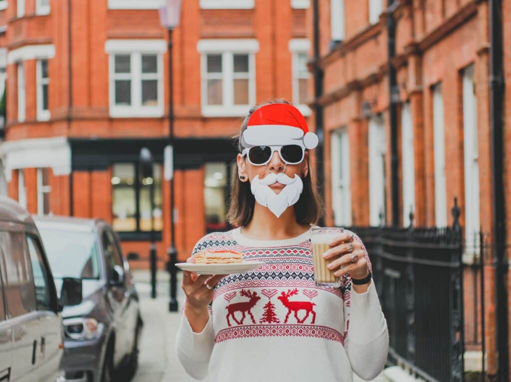 A person wearing a Santa hat, fake white beard, and sunglasses stands outside red brick buildings, holding biscuits and milk in a festive reindeer jumper—serving up fun ideas to update your Christmas family dinner with playful holiday spirit.