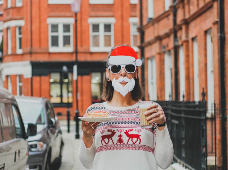 A person wearing a Santa hat, fake white beard, and sunglasses stands outside red brick buildings, holding biscuits and milk in a festive reindeer jumper—serving up fun ideas to update your Christmas family dinner with playful holiday spirit.