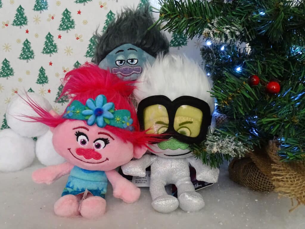 Three colourful Trolls plush toys sit next to a small, decorated Christmas tree with lights. This festive setup with cheerful wallpaper and snowballs makes perfect Christmas Gift Ideas for Children 2022.