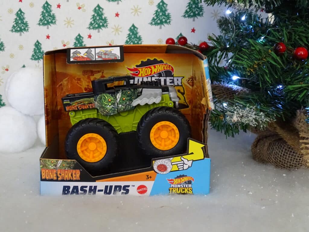 A packaged Hot Wheels Monster Trucks Bone Shaker toy sits on a surface next to a decorated Christmas tree, making it one of the top Christmas Gift Ideas for Children 2022 with its festive background of snow and green stars.