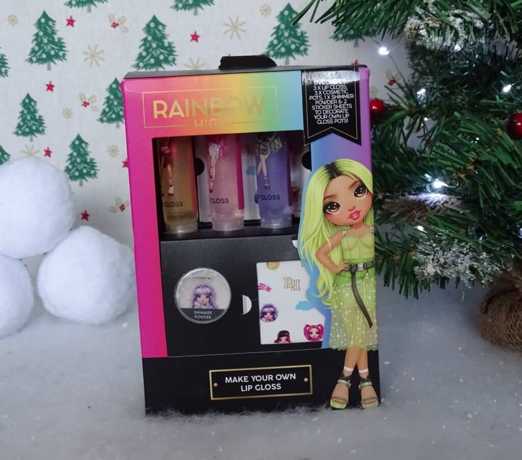 A Rainbow High "Make Your Own Lip Gloss" kit, perfect for Christmas Gift Ideas for Children 2022, is displayed beside a decorated tree and snowball props. The kit features vibrant packaging with an illustrated girl in a green dress on the front.