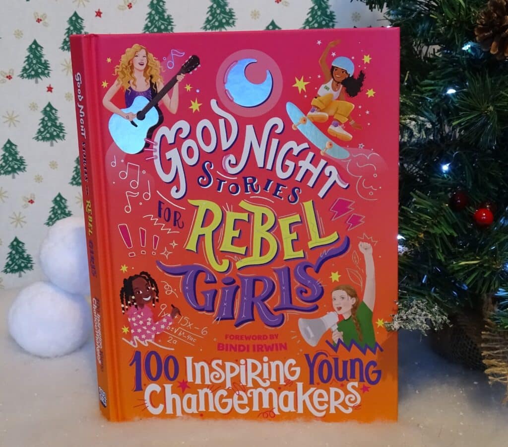 A colourful book titled "Good Night Stories for Rebel Girls: 100 Inspiring Young Changemakers" stands upright beside a decorated Christmas tree, offering perfect Christmas Gift Ideas for Children 2022, with fake snowballs and a festive patterned background.