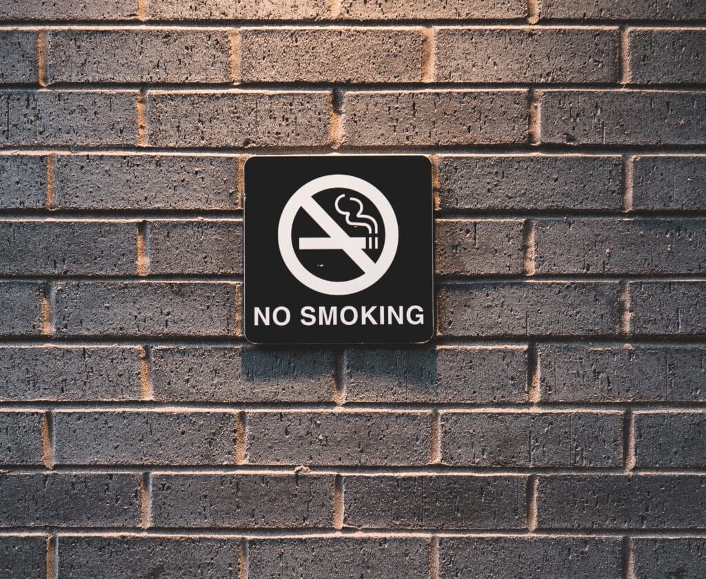 A black and white "No Smoking" sign with a crossed-out cigarette icon is mounted on a grey brick wall, offering a visual reminder for those seeking Five Tips To Help Quit Smoking In 2023.