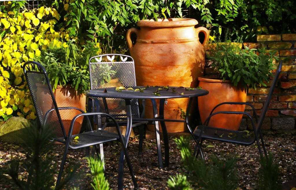 A sunlit garden scene with a round metal table and three mesh chairs, surrounded by lush green plants and large terracotta pots against a brick wall. Fallen leaves are scattered across the garden table and ground.