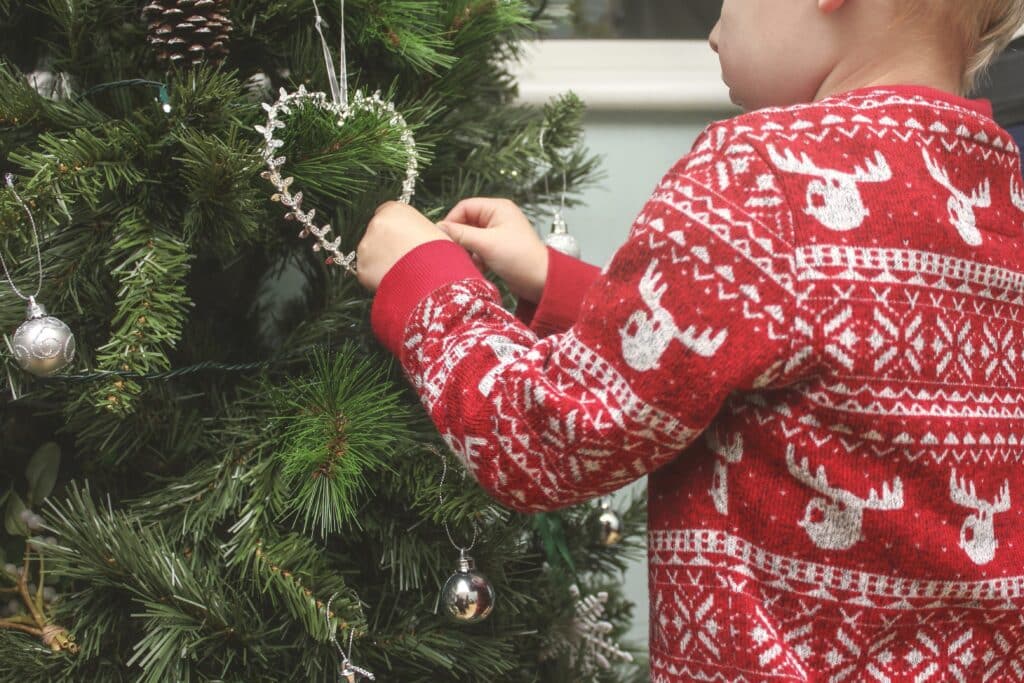 A child in a red jumper with white reindeer patterns hangs a heart-shaped ornament on a decorated Christmas tree—one of many activities to keep children entertained during the Christmas holidays.