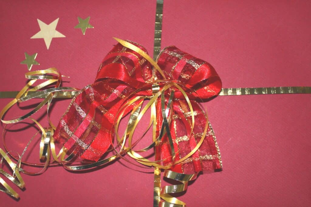 A gift wrapped in red paper with gold ribbon, decorated with a red and gold bow, gold curly ribbons, several gold star-shaped stickers on the top left, and containing a £50 Amazon gift card inside.