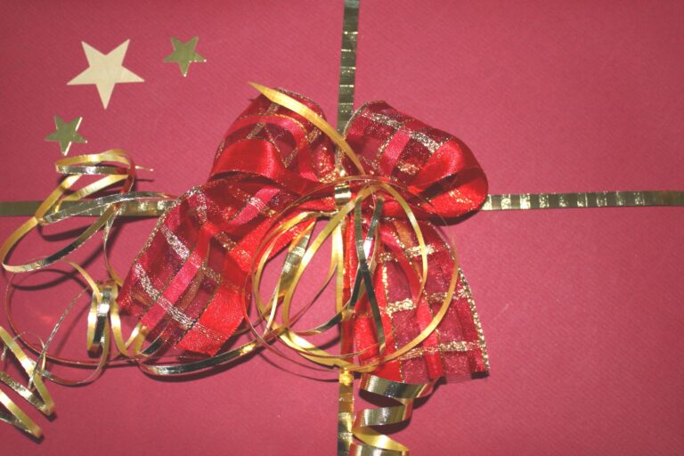 A gift wrapped in red paper with gold ribbon, decorated with a red and gold bow, gold curly ribbons, several gold star-shaped stickers on the top left, and containing a £50 Amazon gift card inside.