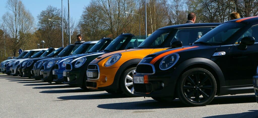 A row of new Mini Coopers in various colours, including orange and blue, parked diagonally in a car park on a sunny day, with a few people standing nearby and trees in the background.