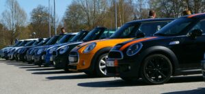 A row of new Mini Coopers in various colours, including orange and blue, parked diagonally in a car park on a sunny day, with a few people standing nearby and trees in the background.