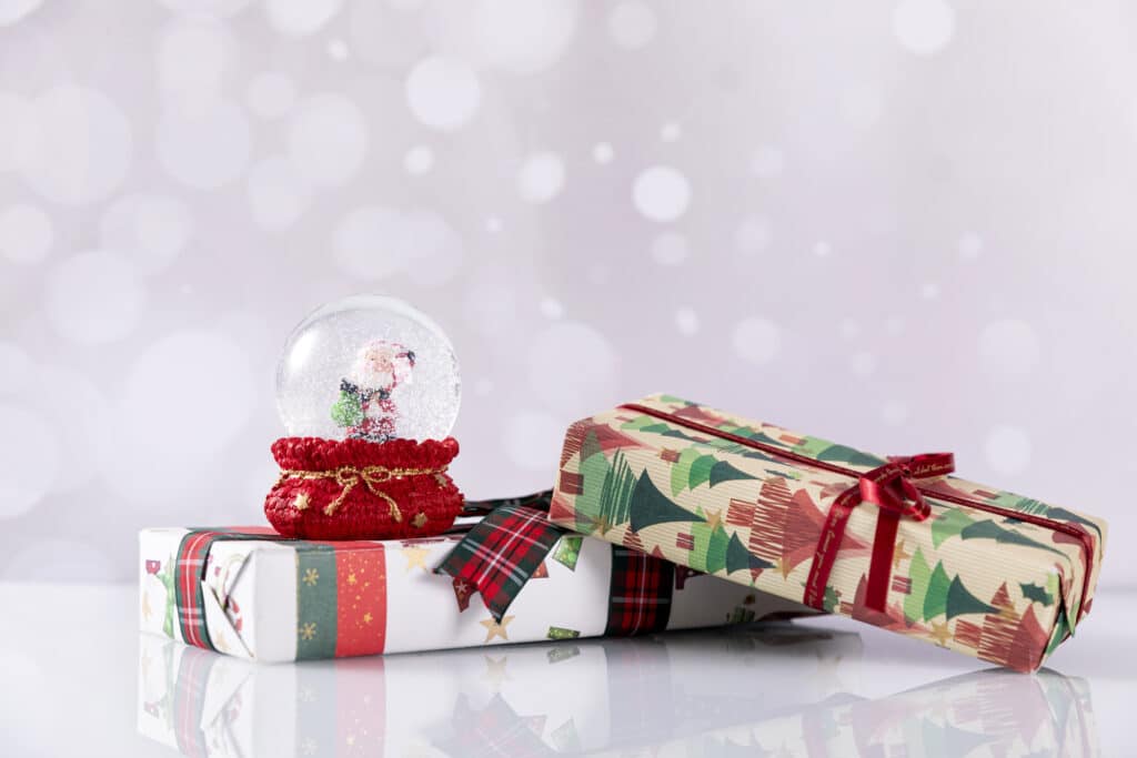 Two wrapped Christmas presents, one with tree patterns and a red ribbon, and the other with stars and green stripes—perfect Christmas Gift Ideas for Children 2022. A Father Christmas-themed snow globe sits on top, framed by blurred white bokeh lights.