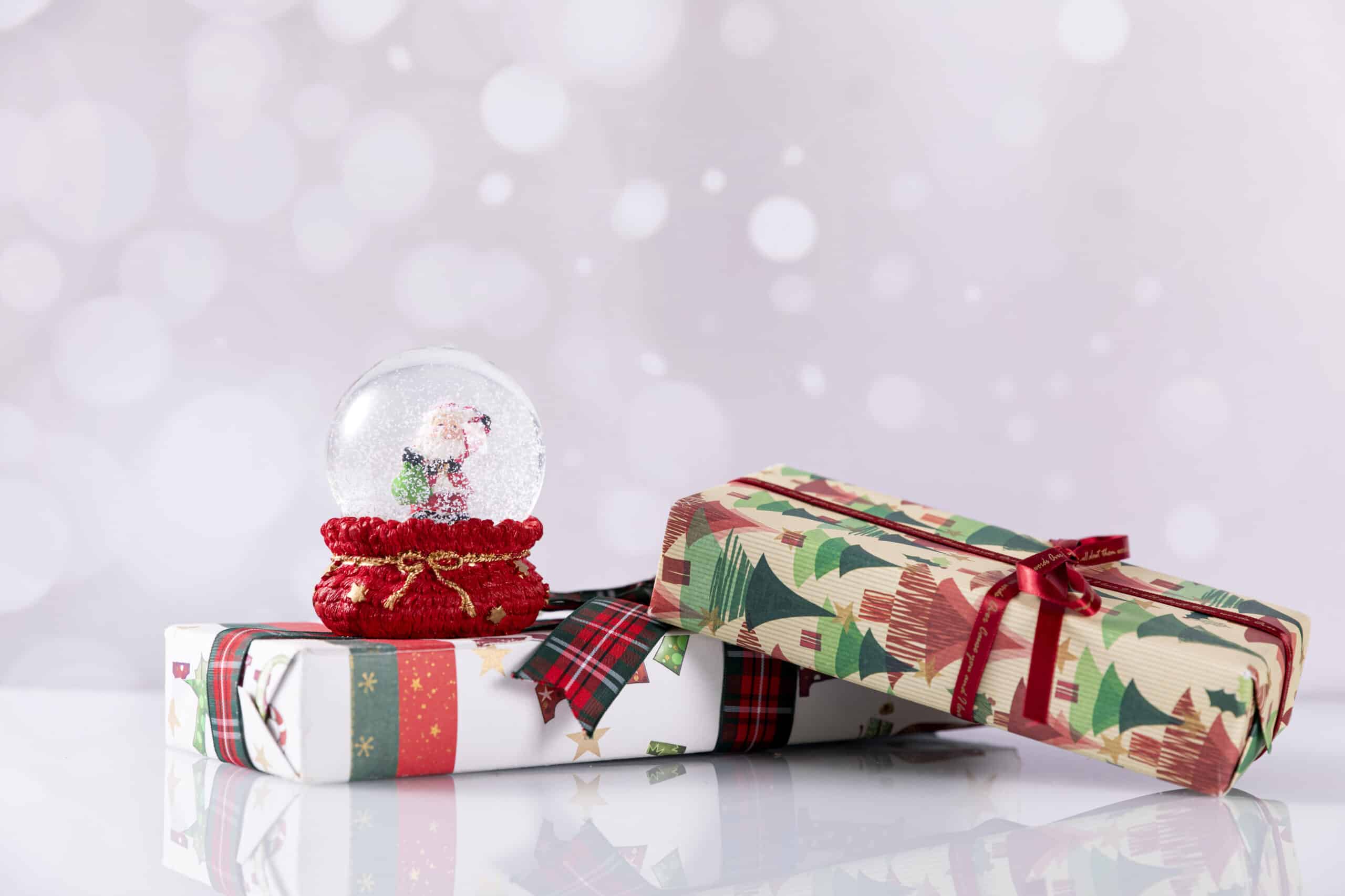 Two wrapped Christmas presents, one with tree patterns and a red ribbon, and the other with stars and green stripes—perfect Christmas Gift Ideas for Children 2022. A Father Christmas-themed snow globe sits on top, framed by blurred white bokeh lights.