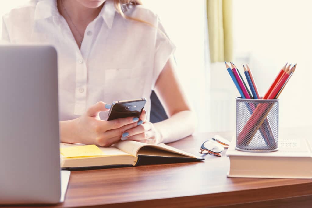A person sits at a desk using a mobile phone, surrounded by an open notebook, closed book, spectacles, a laptop, and coloured pencils—showcasing 3 Must-Know Tips for Creating Your Perfect Office. The person wears a white shirt.