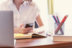 A person sits at a desk using a mobile phone, surrounded by an open notebook, closed book, spectacles, a laptop, and coloured pencils—showcasing 3 Must-Know Tips for Creating Your Perfect Office. The person wears a white shirt.