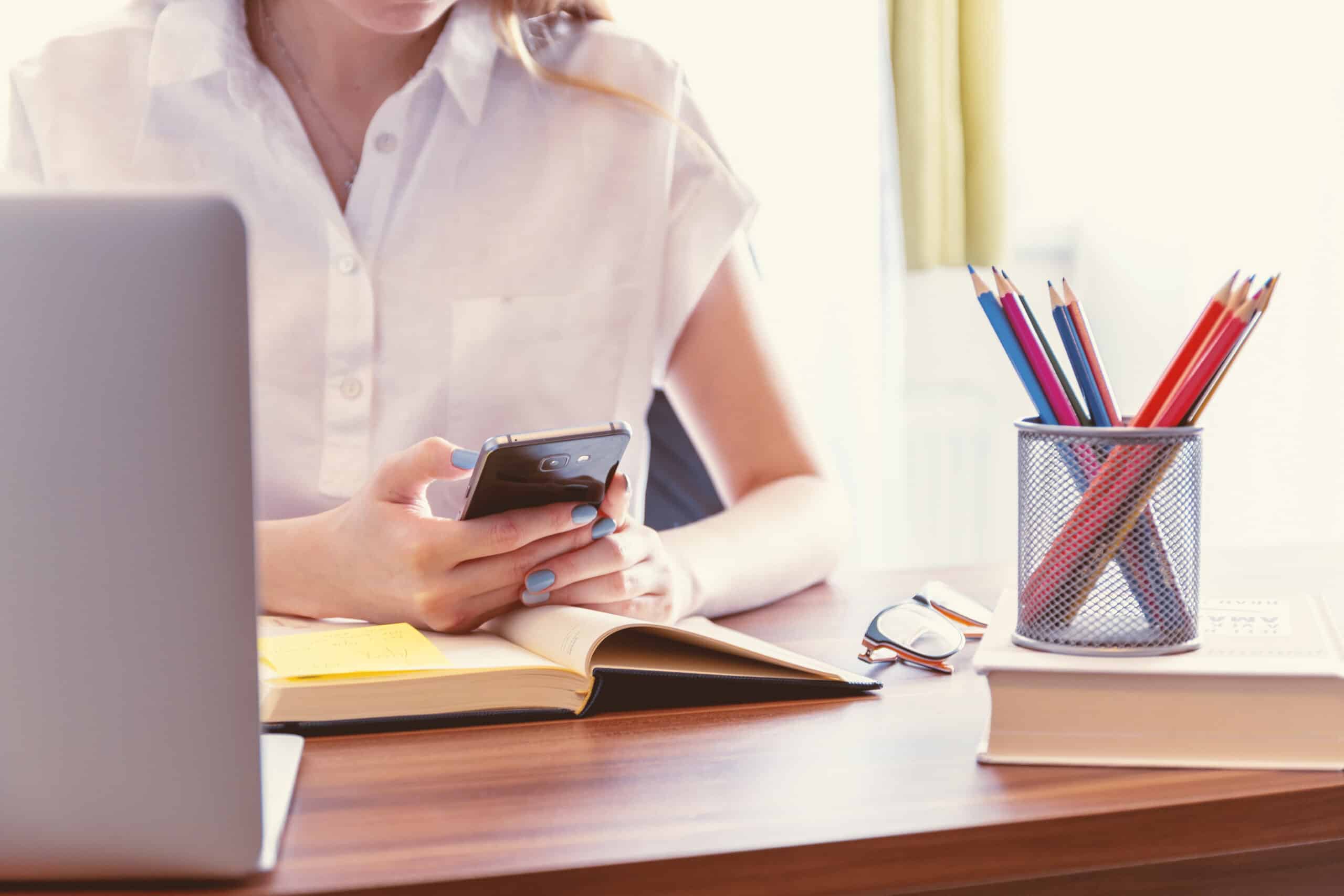 A person sits at a desk using a mobile phone, surrounded by an open notebook, closed book, spectacles, a laptop, and coloured pencils—showcasing 3 Must-Know Tips for Creating Your Perfect Office. The person wears a white shirt.