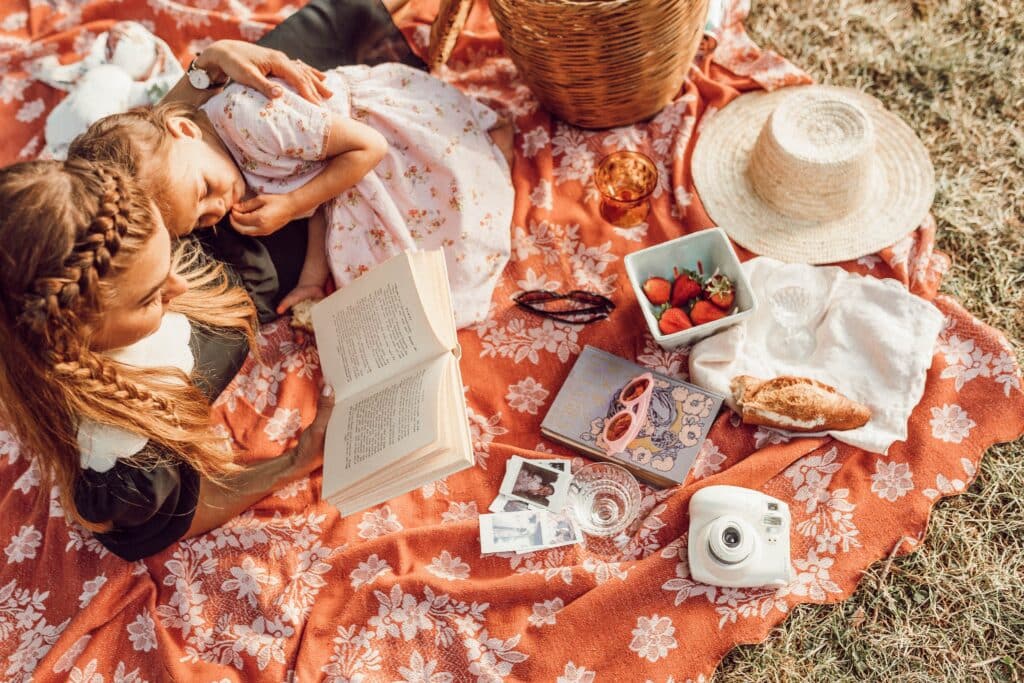 A woman reads a book to a young girl as they relax on a floral blanket during a picnic, sharing quality time together—one of the perfect Mother and Child Date Ideas For Any Budget. Strawberries, Polaroids, and laughter complete the scene.