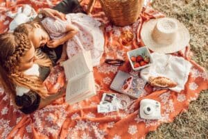 A woman reads a book to a young girl as they relax on a floral blanket during a picnic, sharing quality time together—one of the perfect Mother and Child Date Ideas For Any Budget. Strawberries, Polaroids, and laughter complete the scene.