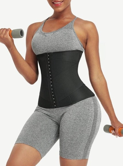 A woman wearing a grey athletic outfit and black waist trainer, showing off one of the fashion investments every woman must have, holds a dumbbell in each hand and smiles against a plain white background.