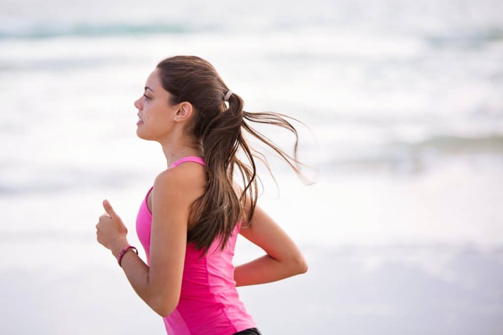 A young woman with long brown hair in a ponytail runs by the sea in a bright pink vest top, embodying 3 Tips to Stay Motivated to Work Out. The blurred, light-coloured background hints at a peaceful beach or waterfront setting.