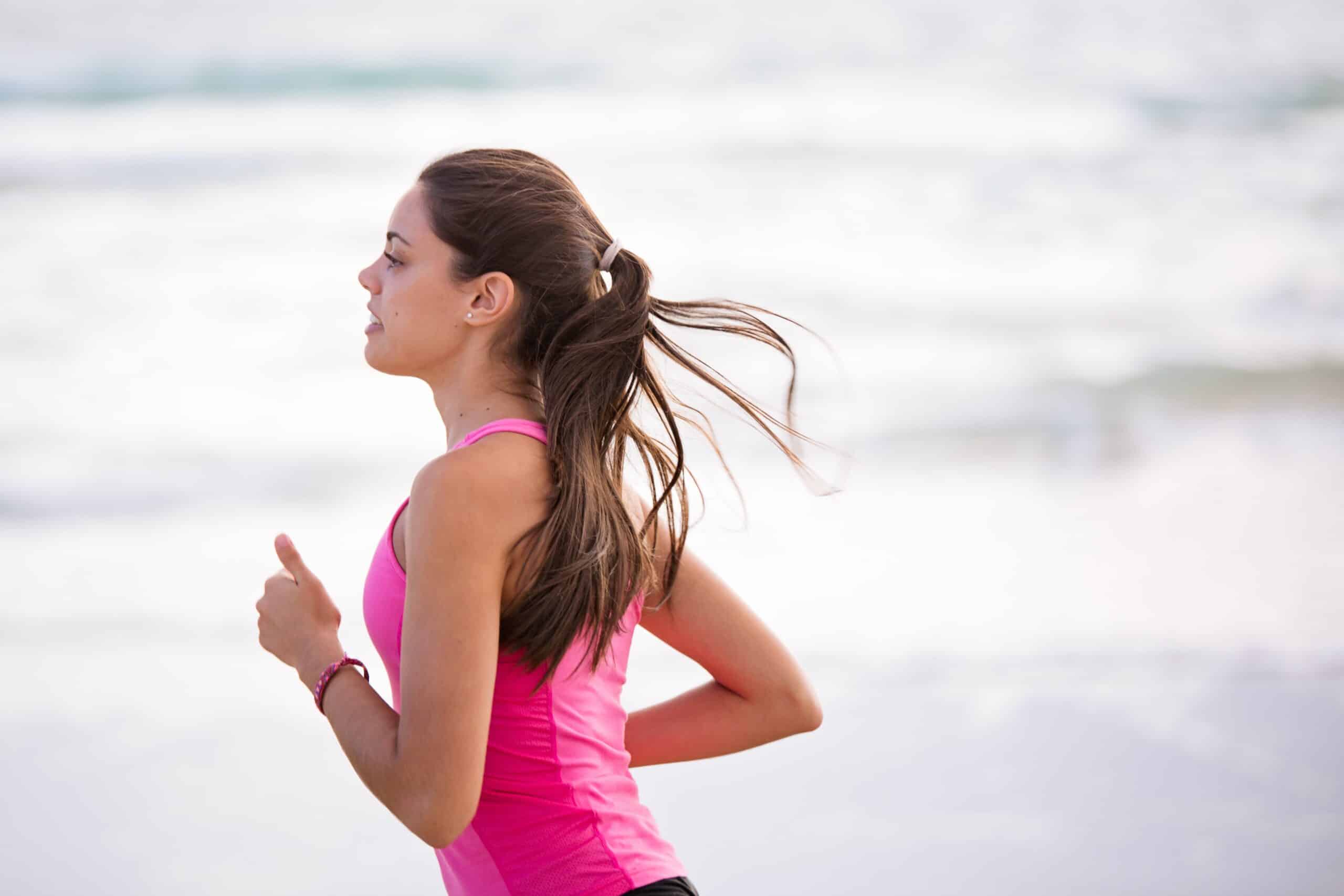 A young woman with long brown hair in a ponytail runs by the sea in a bright pink vest top, embodying 3 Tips to Stay Motivated to Work Out. The blurred, light-coloured background hints at a peaceful beach or waterfront setting.
