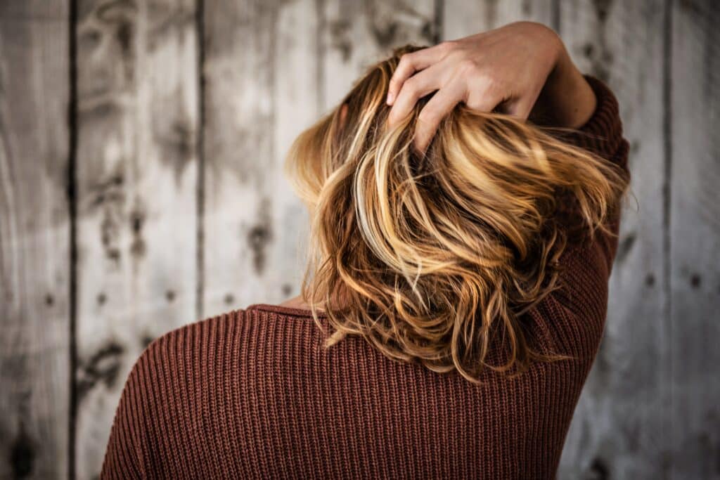 A person with shoulder-length, wavy, blonde hair wearing a brown jumper, seen from behind, holds their head with one hand against a rustic wooden background—prompting questions like: Is hair restoration surgery safe?.