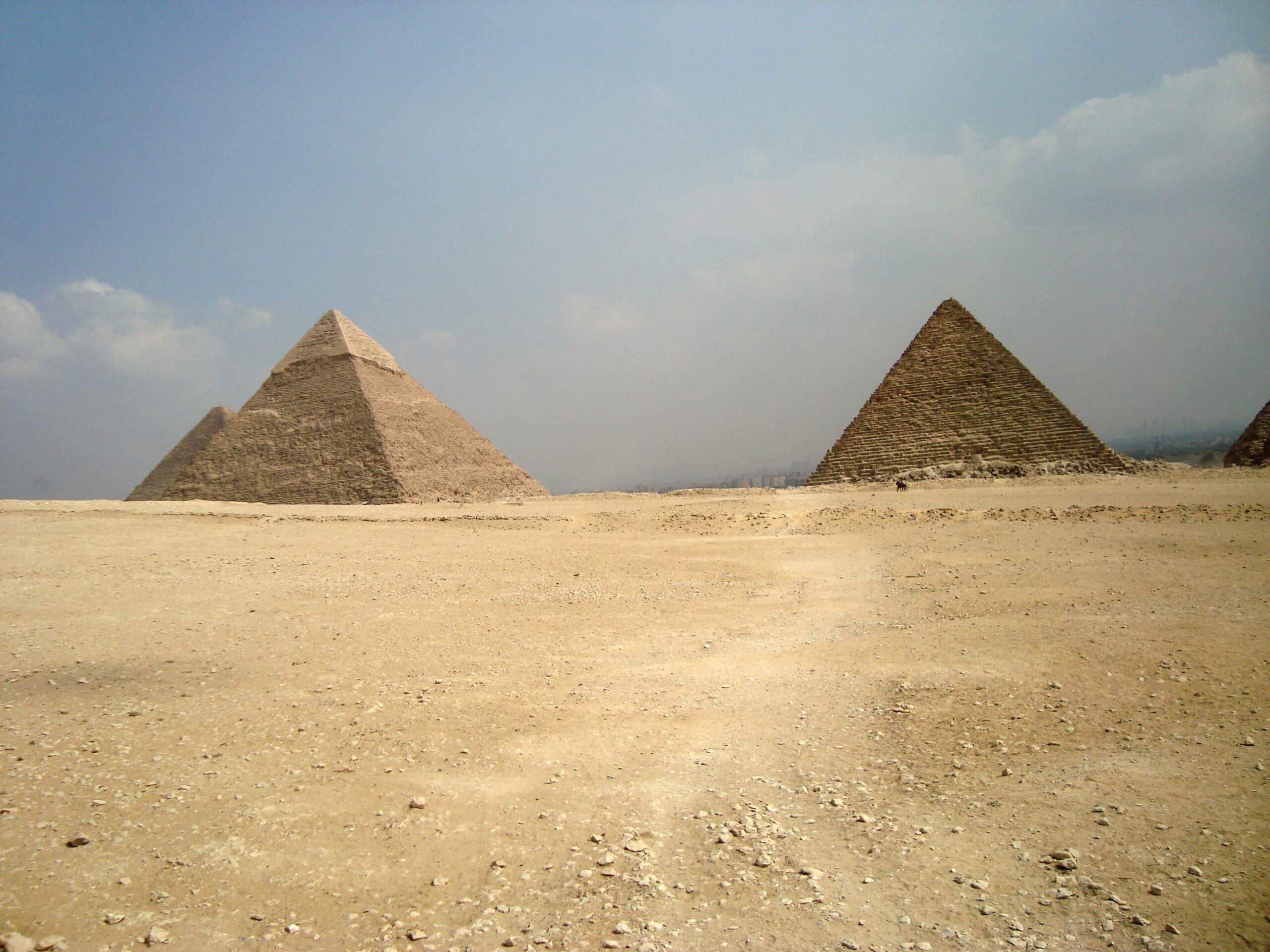Two large pyramids stand in a vast, sandy desert under a hazy, partly cloudy sky. With their smooth sides and rocky surroundings, you might wonder: Are The Pyramids Really Worth Adding To Your Travel Bucket List?.