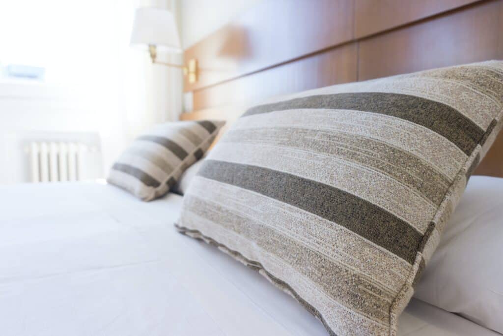 Two large, beige and grey striped pillows rest on a neatly made bed with white sheets in a bright, sunlit room—a welcoming touch when learning how to create the perfect guest bedroom. A lamp and window are visible in the background.