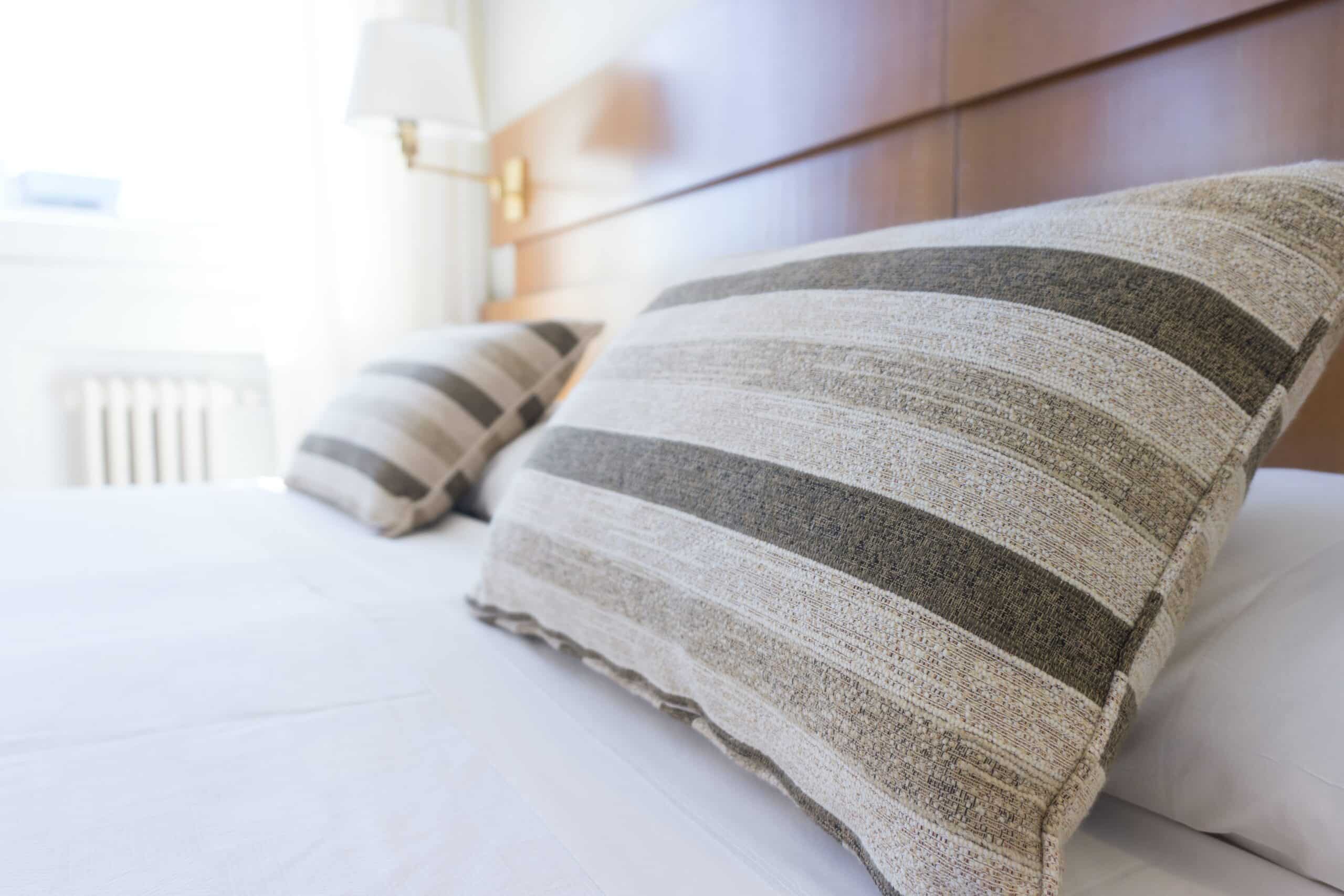 Two large, beige and grey striped pillows rest on a neatly made bed with white sheets in a bright, sunlit room—a welcoming touch when learning how to create the perfect guest bedroom. A lamp and window are visible in the background.