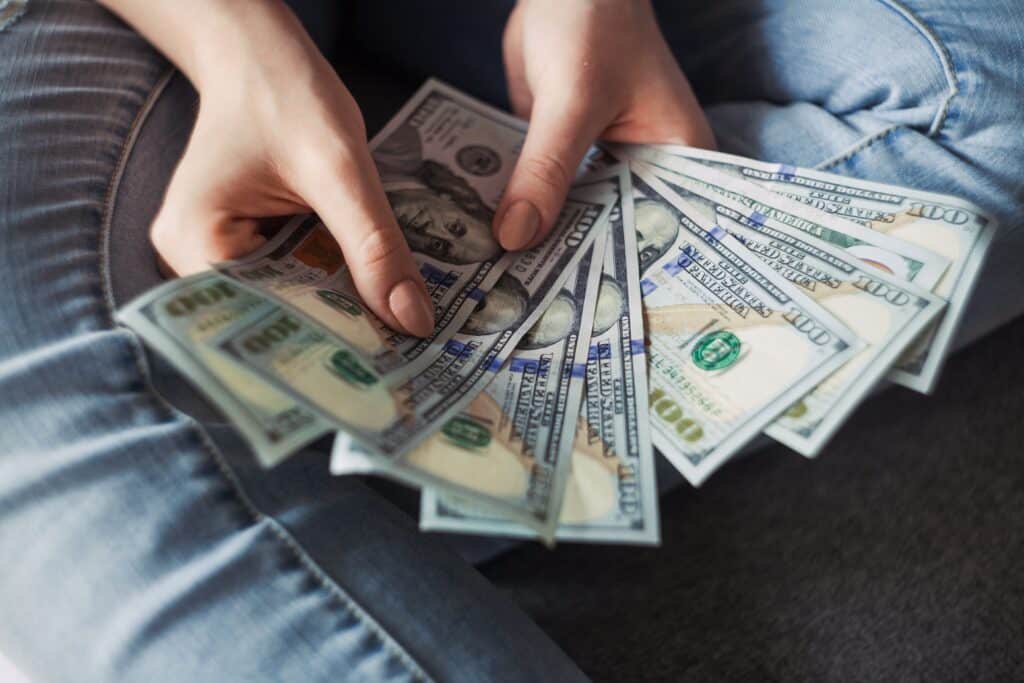 A person wearing blue jeans is holding and spreading out several US $100 notes in their hands, showing the money clearly—ready to Save Money this Winter.