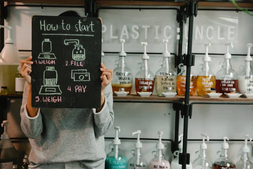A person holds a chalkboard with illustrated refill instructions—1. Weigh, 2. Fill, 3. Weigh, 4. Pay—in front of shelves lined with reusable bottles and liquid dispensers to help Increase Sustainability in Your Business at a zero-waste shop.