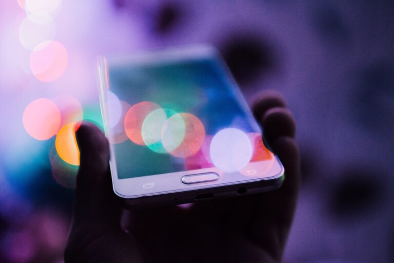 A hand holds a mobile phone with a blurred, colourful background of bokeh lights, evoking a dreamy and abstract atmosphere—perfect for reaching out to Christmas Helplines during the festive season. The phone’s screen reflects the vibrant light effects.