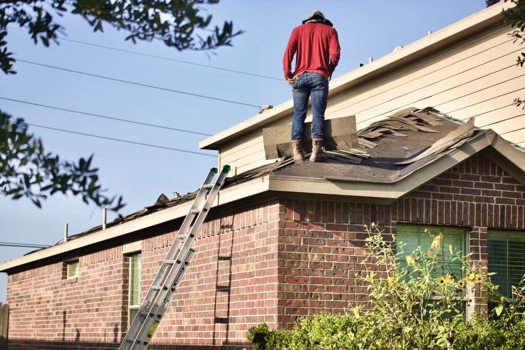 A person in a red hoodie and jeans stands on a house roof under repair, with tiles removed—a reminder of the importance of the criteria used in selecting the best roofing company. A ladder leans against the brick wall, framed by trees on a sunny day.
