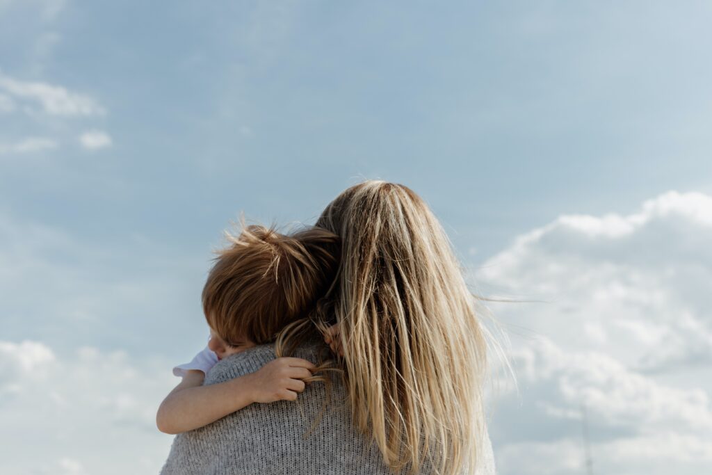A woman with long blonde hair holds a small child with brown hair in her arms, both facing away from the camera, as they share lessons in emotional development beneath a bright blue sky with scattered white clouds.
