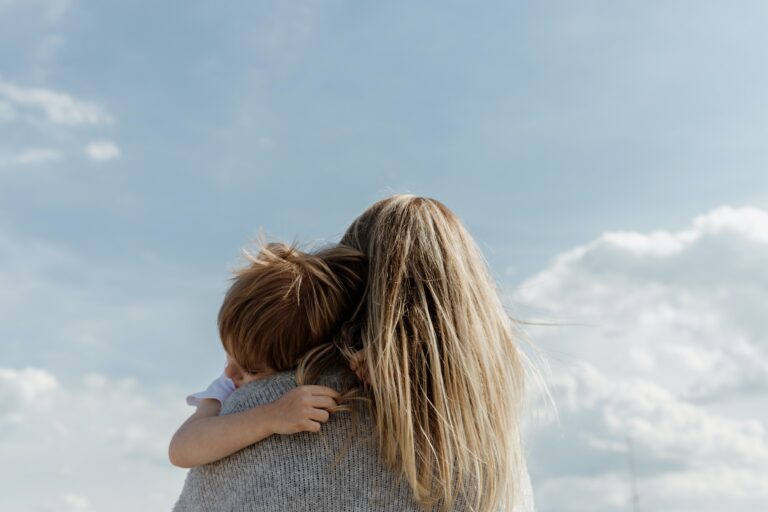 A woman with long blonde hair holds a small child with brown hair in her arms, both facing away from the camera, as they share lessons in emotional development beneath a bright blue sky with scattered white clouds.