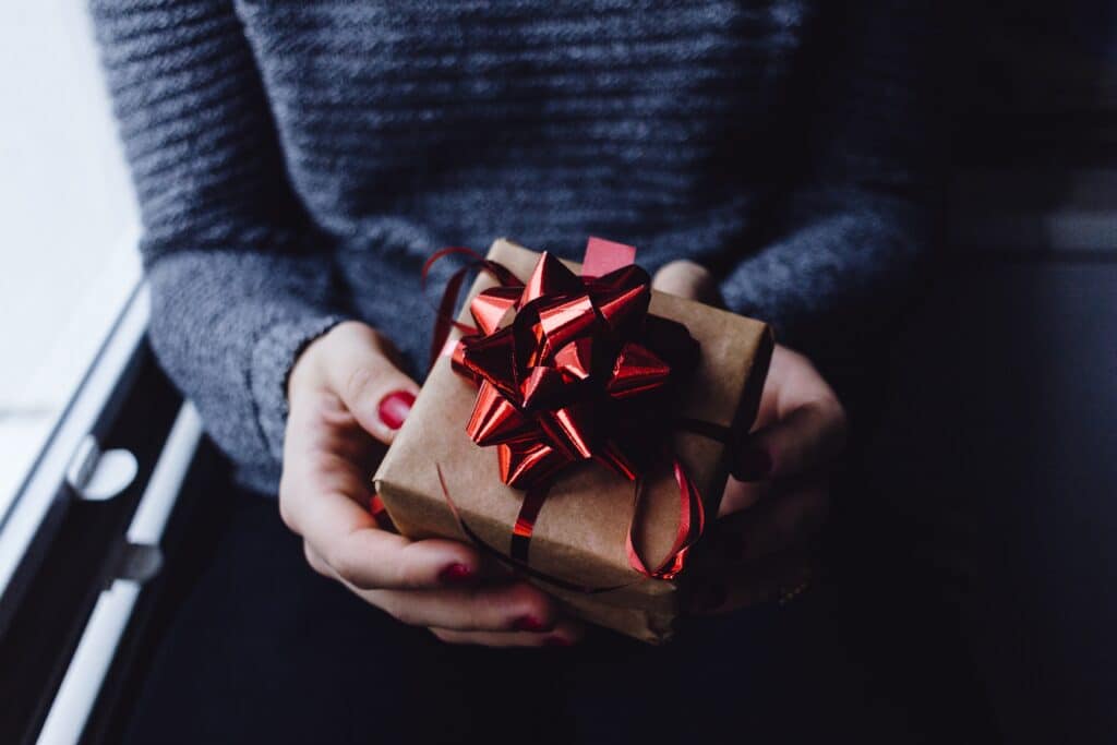 A person wearing a grey jumper and red nail varnish holds a small present wrapped in brown paper with a shiny red bow—perfect inspiration for 5 Gift Ideas Everyone Will Love. The softly lit background adds to the cosy atmosphere.