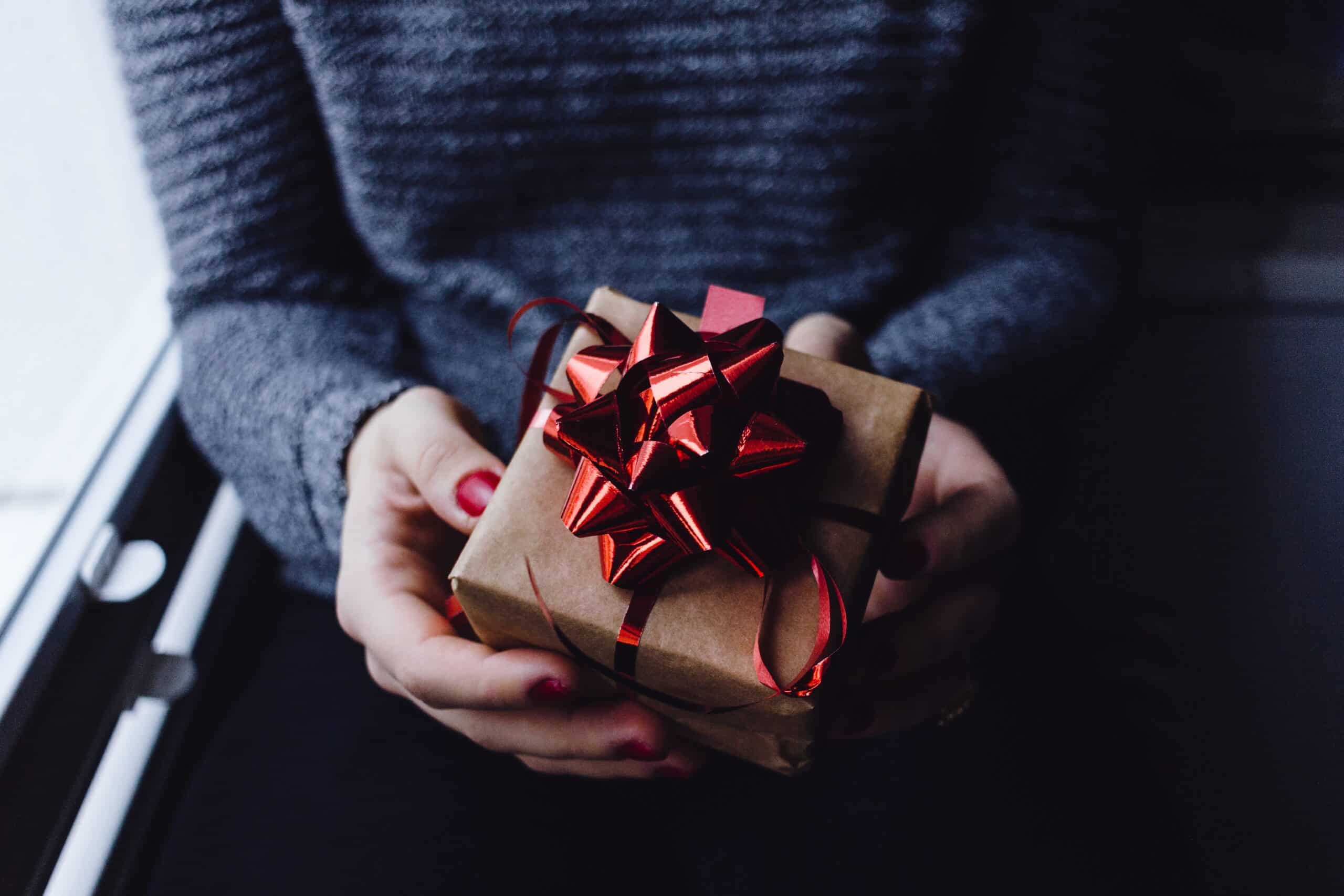 A person wearing a grey jumper and red nail varnish holds a small present wrapped in brown paper with a shiny red bow—perfect inspiration for 5 Gift Ideas Everyone Will Love. The softly lit background adds to the cosy atmosphere.