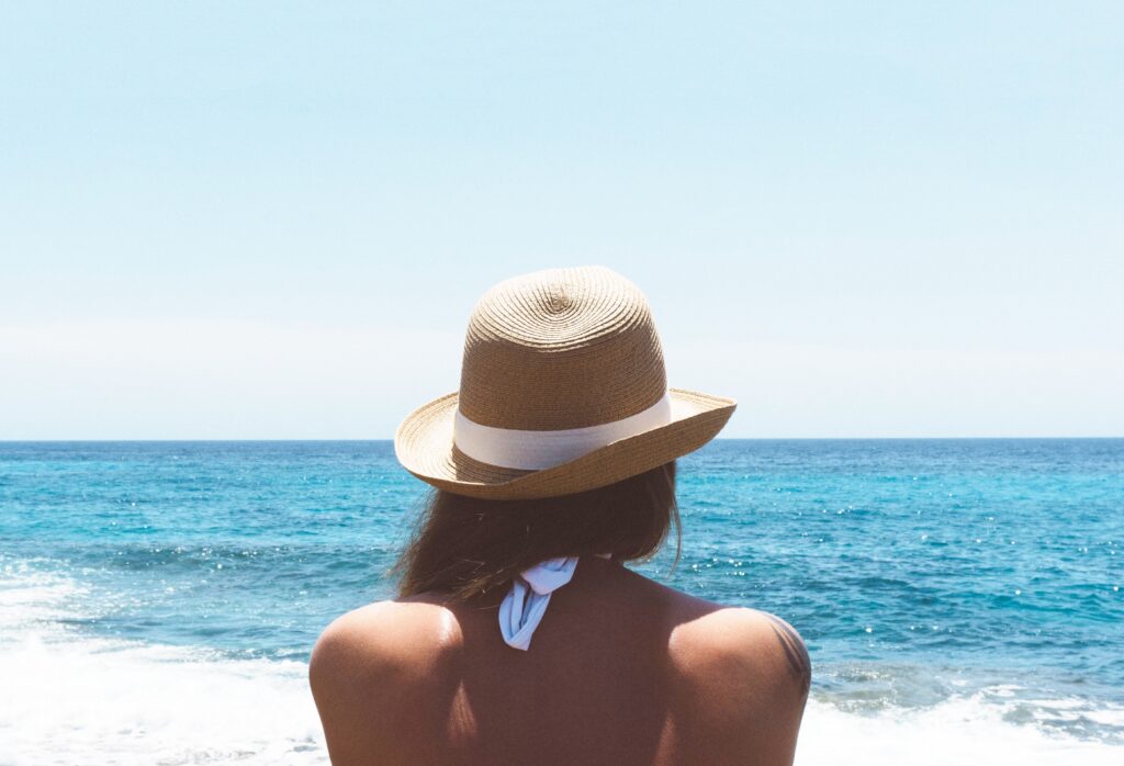 A person wearing a straw hat and white swimming costume looks out at the blue sea under a clear sky. This serene moment prompts reflection: How Is Clothing Related To Women's Empowerment? Choices in attire can express confidence and individuality.