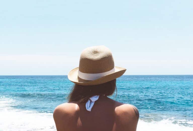 A person wearing a straw hat and white swimming costume looks out at the blue sea under a clear sky. This serene moment prompts reflection: How Is Clothing Related To Women's Empowerment? Choices in attire can express confidence and individuality.