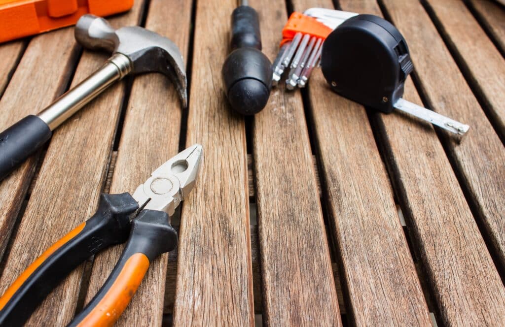 A hammer, pliers, screwdriver with interchangeable bits, and a tape measure are arranged on a wooden surface—essentials for any DIY or repair project. These tools showcase practical tips to stay on top of home maintenance.