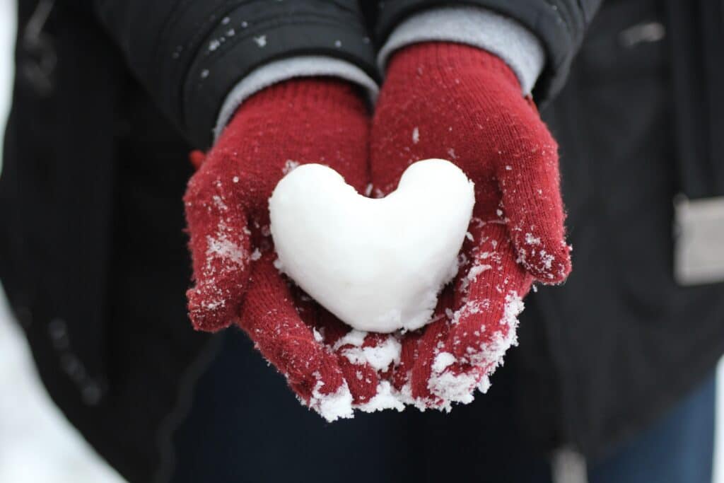 A person wearing red gloves and winter clothing holds a heart-shaped snowball in their hands, with snow dusting the gloves and a dark jacket and blue trousers visible in the background.