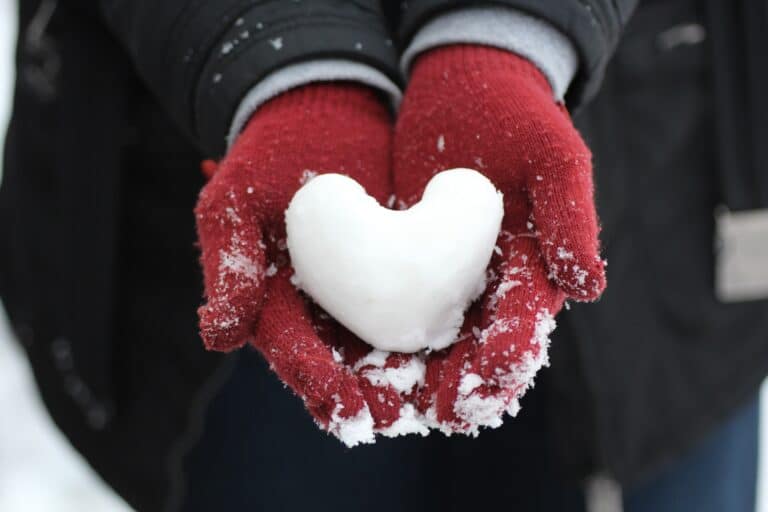 A person wearing red gloves and winter clothing holds a heart-shaped snowball in their hands, with snow dusting the gloves and a dark jacket and blue trousers visible in the background.