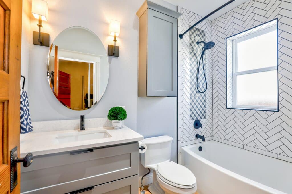 Modern bathroom with grey vanity, oval mirror, two wall lights, white worktop, potted plant, toilet, and bath with black fittings. The shower wall features a white herringbone tile pattern and window—perfect for luxury bathroom refurbishments.