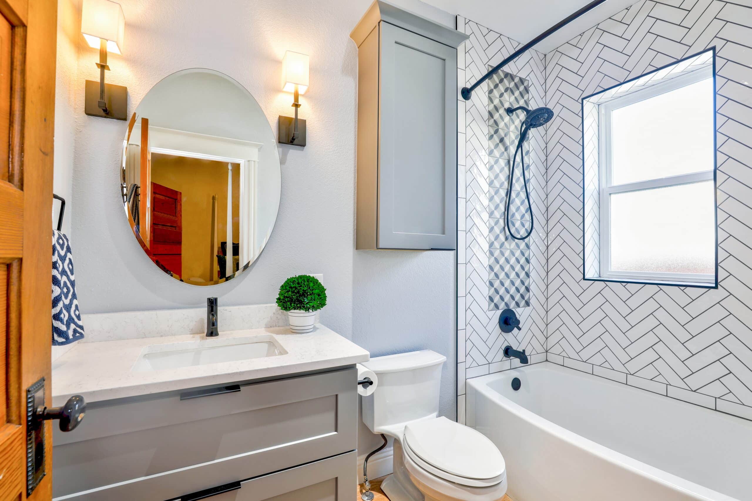 Modern bathroom with grey vanity, oval mirror, two wall lights, white worktop, potted plant, toilet, and bath with black fittings. The shower wall features a white herringbone tile pattern and window—perfect for luxury bathroom refurbishments.