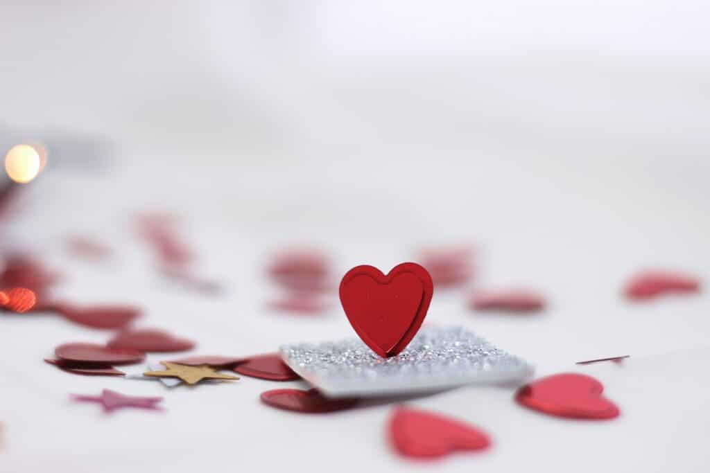 A small red heart stands upright on a silver glittery square, surrounded by Valentine’s Day-themed red and gold heart- and star-shaped confetti on a white surface.