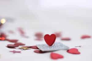 A small red heart stands upright on a silver glittery square, surrounded by Valentine’s Day-themed red and gold heart- and star-shaped confetti on a white surface.