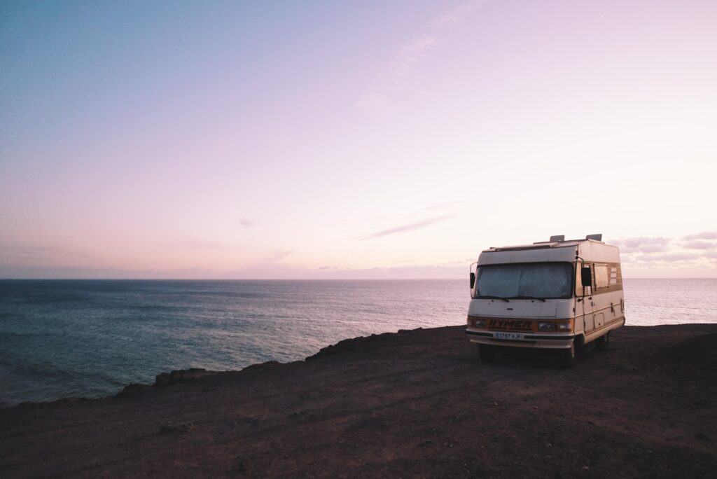 A white campervan is parked on a rocky cliff overlooking a calm sea at sunset, with a pink and purple sky in the background—perfect inspiration for tips for planning a fantastic road trip.