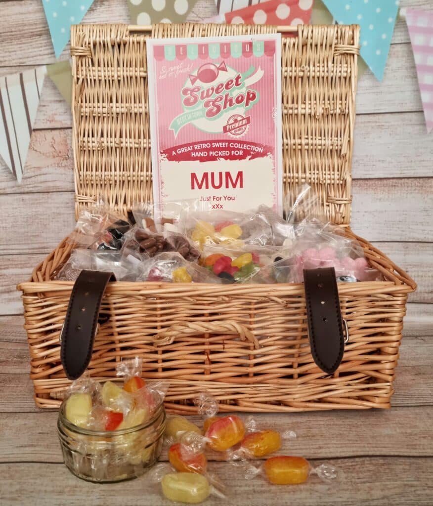 A wicker hamper filled with assorted retro sweets in clear wrappers sits open on a wooden surface. A sign reads “Sweet Shop” and “Hand picked for Mum.” Celebrate Mother’s Day with Very—sweet treats and pastel bunting set the scene.