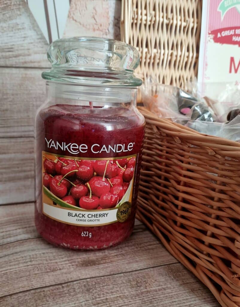 A large glass jar of Yankee Candle in the Black Cherry scent sits next to a wicker basket on a wooden surface. Perfect for celebrating Mother’s Day with Very, the label shows ripe cherries and the jar holds deep red wax.