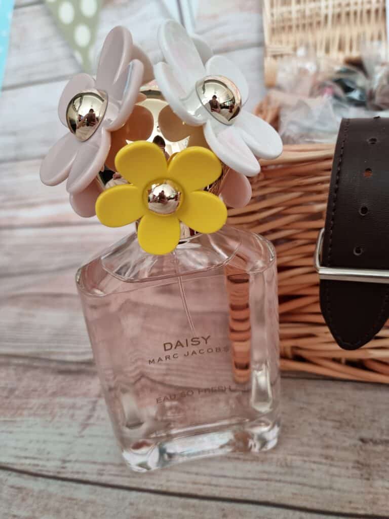A bottle of Daisy Marc Jacobs perfume with a floral cap, featuring white and yellow flower designs, sits on a wooden surface next to a woven basket and a dark brown belt—a perfect Mother’s Day with Very gift.